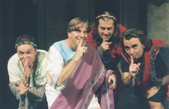 Four costumed performers lean together and hold fingers to their lips in a shushing gesture.
