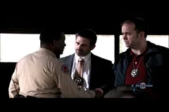 Three men, two wearing badges, confer inside a dimly lit vehicle.
